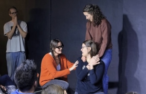 Performers on a small stage engaged in an emotional scene, with one person kneeling and others offering comfort as an audience watches.
