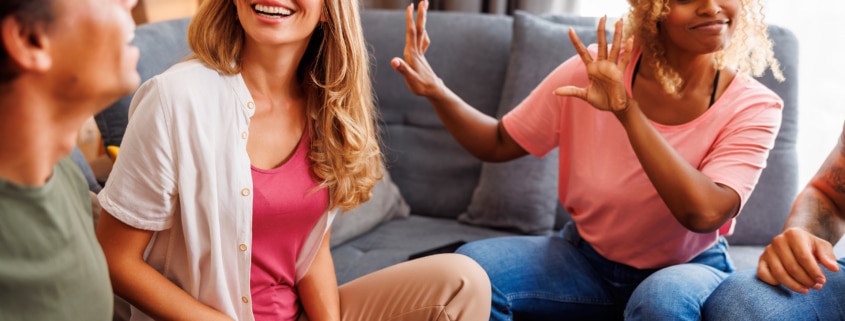 Group of friends sitting together on a couch, actively gesturing and laughing while playing charades indoors.