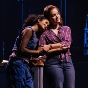 Two performers onstage, with one leaning in close to speak to the other beside a kitchen counter during a dramatic scene.