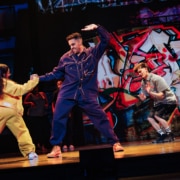 A group of dancers performing an energetic choreography onstage, framed by a colorful graffiti backdrop.