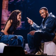 Two performers seated on low stools during an emotional onstage conversation, with one gesturing as they talk.