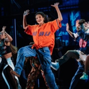 A large ensemble of dancers mid-choreography, arms raised and moving in sync during a high-energy musical number.