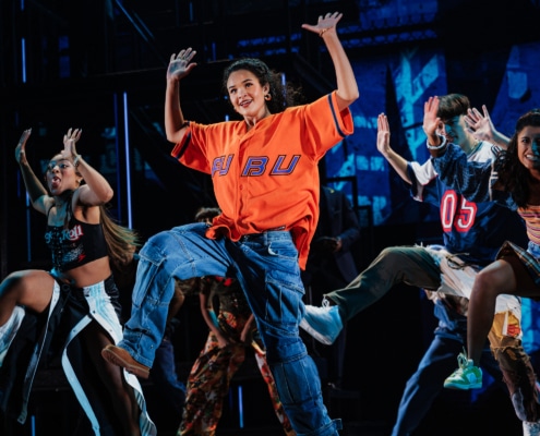 A large ensemble of dancers mid-choreography, arms raised and moving in sync during a high-energy musical number.
