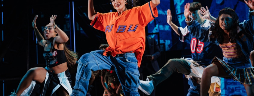 A large ensemble of dancers mid-choreography, arms raised and moving in sync during a high-energy musical number.