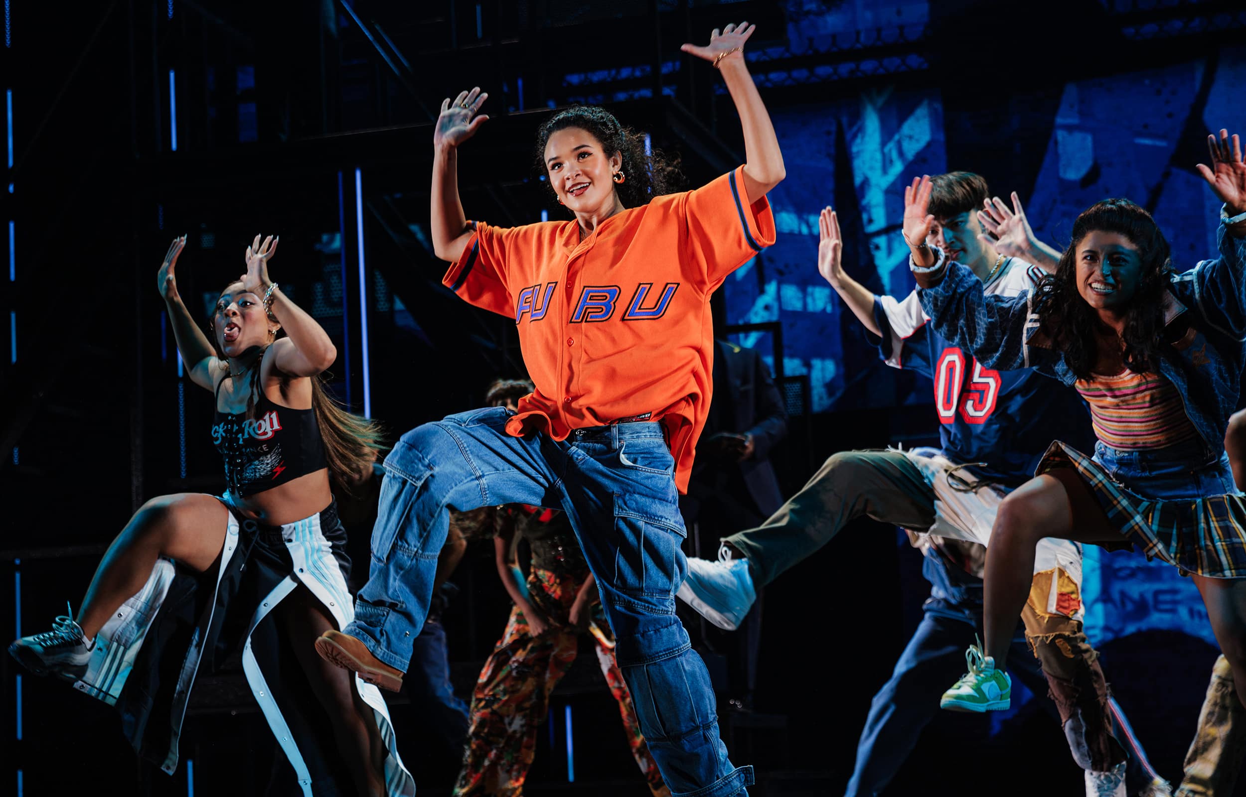 A large ensemble of dancers mid-choreography, arms raised and moving in sync during a high-energy musical number.