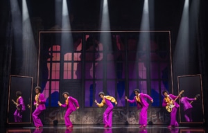MJ musical performer leading a dynamic group dance scene in a rehearsal‑style setting with full ensemble on stage