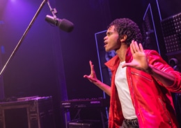 Performer in a red jacket stands onstage near a microphone, holding both hands up in a studio‑style setting with musical equipment in the background.