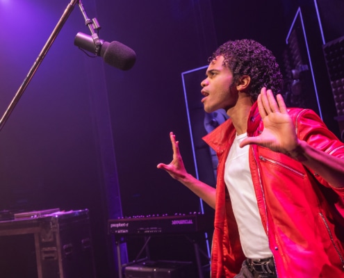 Performer in a red jacket stands onstage near a microphone, holding both hands up in a studio‑style setting with musical equipment in the background.
