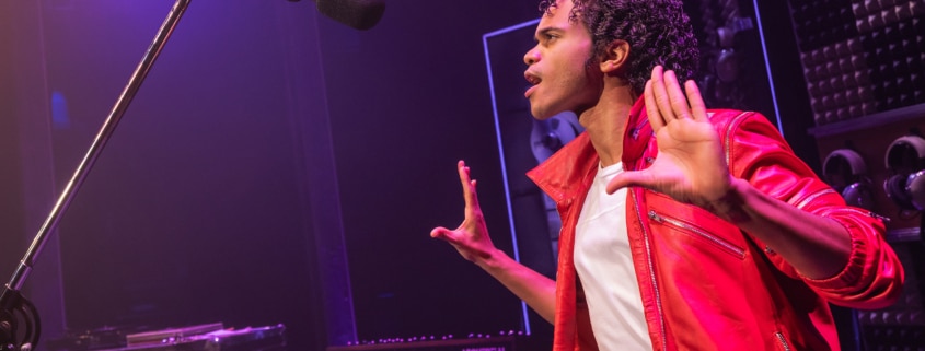 Performer in a red jacket stands onstage near a microphone, holding both hands up in a studio‑style setting with musical equipment in the background.