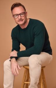 Person seated on a wooden stool wearing a dark green sweater and light beige pants against a warm tan studio backdrop.