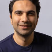 A person with dark hair wearing a navy blue shirt with a small embroidered logo, photographed against a plain light gray background