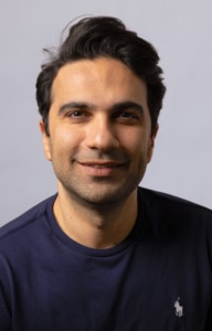 A person with dark hair wearing a navy blue shirt with a small embroidered logo, photographed against a plain light gray background