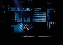 Theatrical stage performance with two performers at a piano in a dimly lit scene featuring a backdrop labeled ‘Ellington Room’.