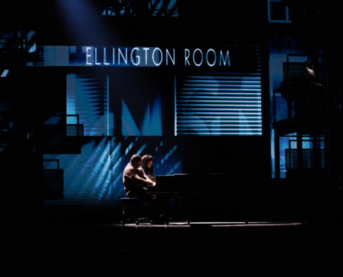 Theatrical stage performance with two performers at a piano in a dimly lit scene featuring a backdrop labeled ‘Ellington Room’.