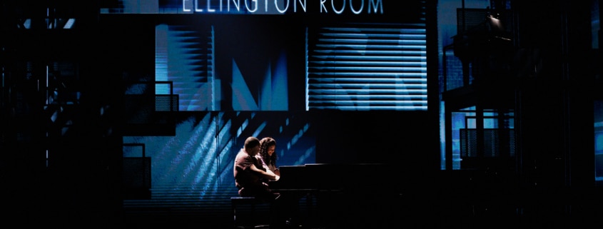 Theatrical stage performance with two performers at a piano in a dimly lit scene featuring a backdrop labeled ‘Ellington Room’.