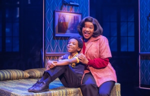 An adult and a child sit together on a bed in a warmly lit room with patterned walls and framed artwork in the background.