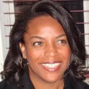 Close-up portrait of a person indoors, shown from the shoulders up, wearing a dark top, with horizontal window blinds in the background.