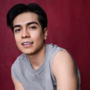 Person in a gray sleeveless shirt posed against a deep red backdrop