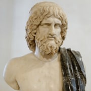 Marble bust of a draped, bearded figure with detailed curly hair, shown from the chest up and wearing a dark-veined marble garment over one shoulder.