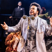 Performer in a light-colored suit sings or speaks into a vintage-style dual microphone, with ensemble members reacting and a band visible on an upper level.