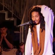 Person standing at a microphone onstage, wearing layered clothing, with several seated people and stair railings visible in the background.