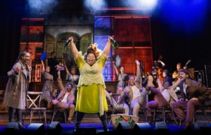 nsemble cast celebrates onstage as a central performer in a bright green dress raises two bottles overhead, surrounded by chairs, props, and warm theatrical lighting.
