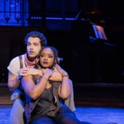 Two performers sit close together on the stage floor, one seated behind the other with arms gently around the front performer, under cool blue lighting.