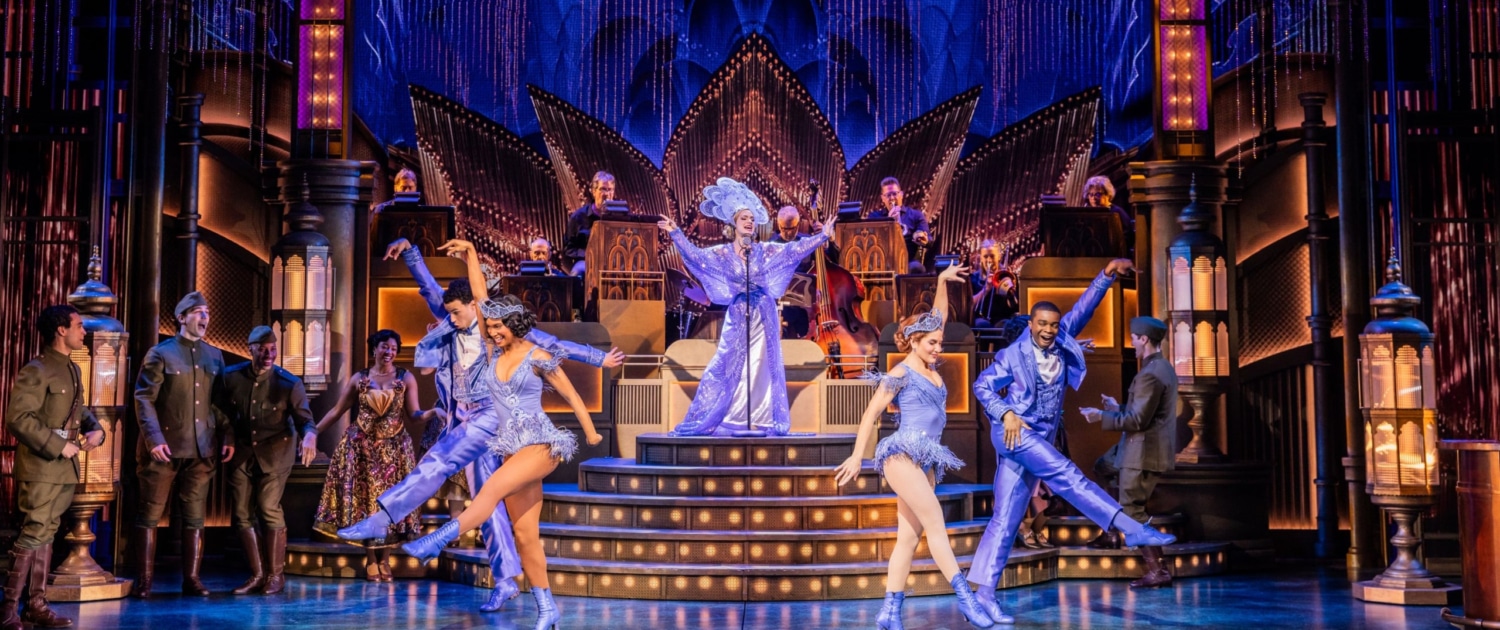 Photo of a lavish stage production with four dancers, one singer, onstage musicians, and individuals in military attire looking on.