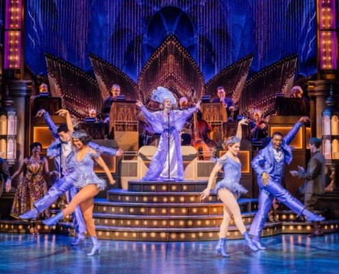 A lavish stage production with 4 dancers, 1 singer, onstage musicians and several actors in military attire looking on.