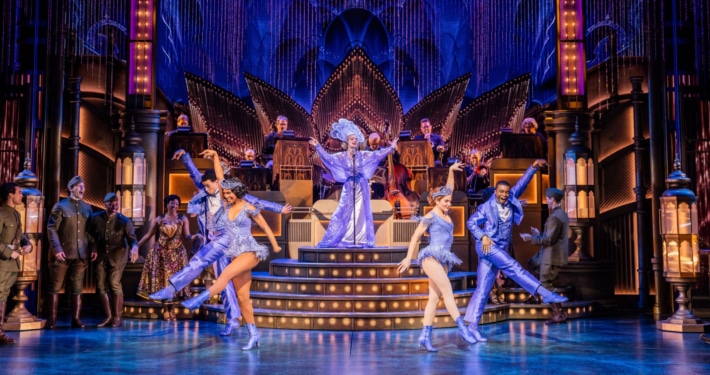 A lavish stage production with 4 dancers, 1 singer, onstage musicians and several actors in military attire looking on.