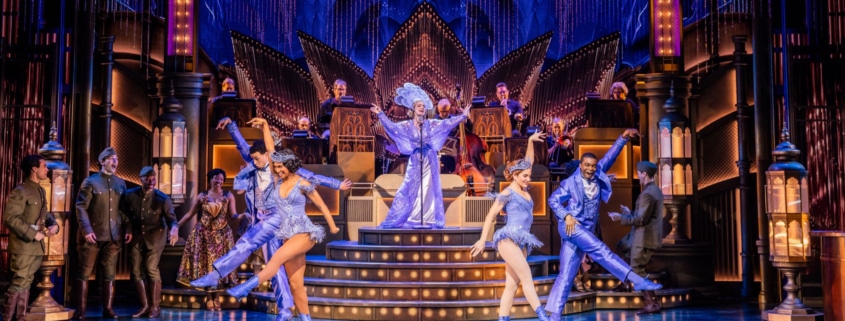 A lavish stage production with 4 dancers, 1 singer, onstage musicians and several actors in military attire looking on.