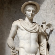 Marble statue of a standing figure wearing a brimmed hat and a draped cloak, holding a staff with wing-like details, shown against a textured stone backdrop.