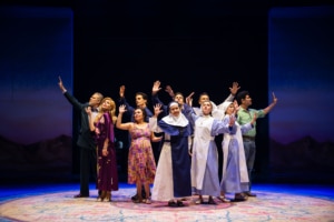 An ensemble cast stands in a semicircle with raised hands on a theater stage, including performers in everyday clothing and nun habits.
