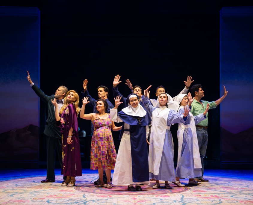 An ensemble cast stands in a semicircle with raised hands on a theater stage, including performers in everyday clothing and nun habits.