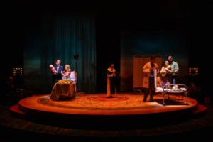 Two family scenes unfold simultaneously onstage: one couple cradles a baby in a hospital bed while another group gathers around a newborn near a bassinet.