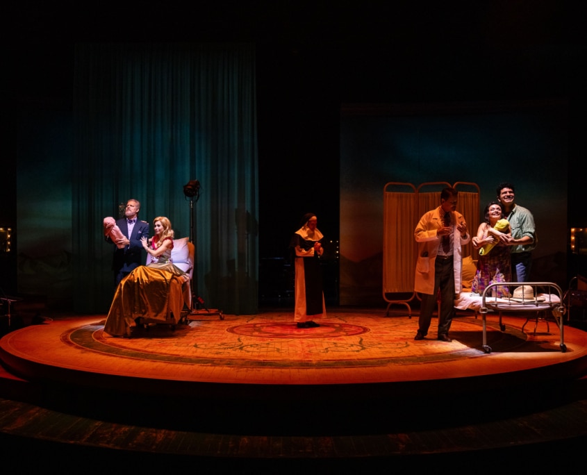 Two family scenes unfold simultaneously onstage: one couple cradles a baby in a hospital bed while another group gathers around a newborn near a bassinet.