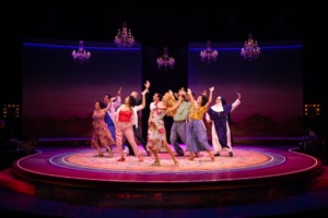 An ensemble dances beneath sparkling chandeliers on a circular stage glowing with pink and purple lighting.
