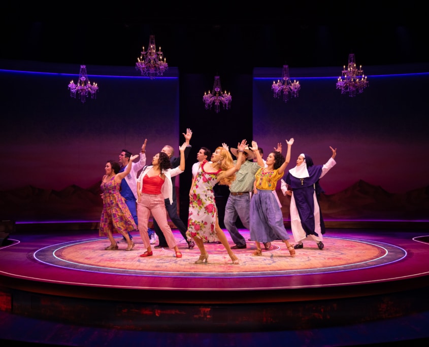 An ensemble dances beneath sparkling chandeliers on a circular stage glowing with pink and purple lighting.
