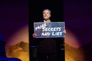 A performer holds a sign reading “MORE SECRETS AND LIES” while standing alone in a spotlight onstage.
