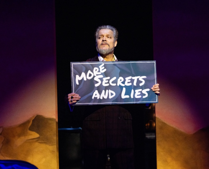 A performer holds a sign reading “MORE SECRETS AND LIES” while standing alone in a spotlight onstage.
