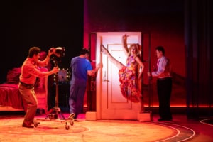A performer poses against a doorway under red lighting while other cast members operate stage lights and interact with the set.