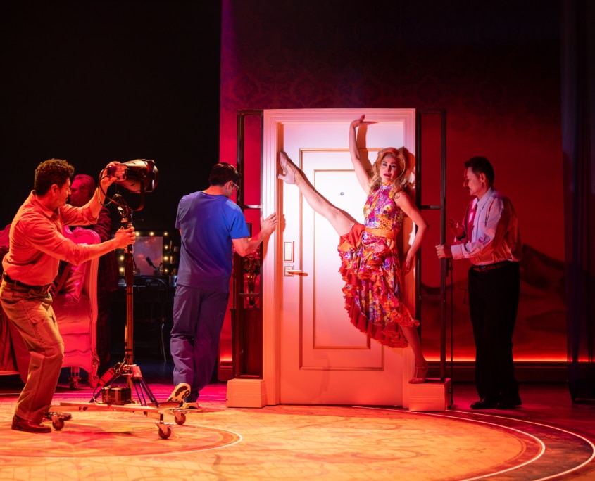 A performer poses against a doorway under red lighting while other cast members operate stage lights and interact with the set.