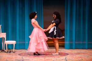 Two performers, one in a pink dress and one in a maids uniform, hold hands at center stage in front of flowing blue curtains.