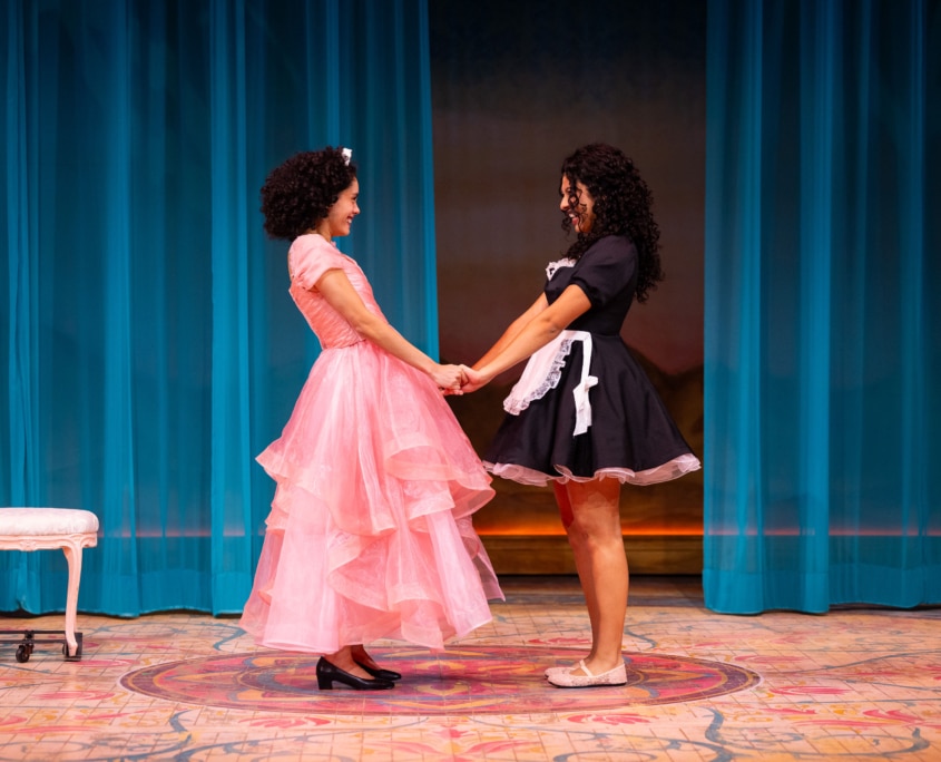 Two performers, one in a pink dress and one in a maids uniform, hold hands at center stage in front of flowing blue curtains.