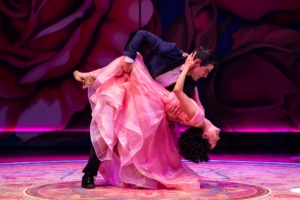 A performer in a pink gown is dipped low in a dance pose by a partner against a backdrop of oversized painted roses.