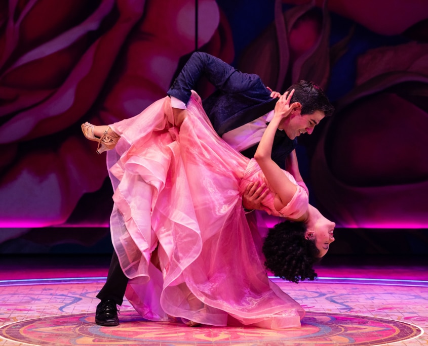 A performer in a pink gown is dipped low in a dance pose by a partner against a backdrop of oversized painted roses.