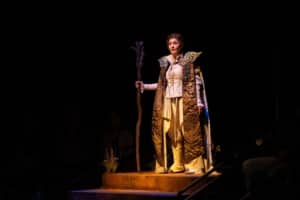 A performer wearing a long, textured robe stands on a raised platform holding a wooden staff under a spotlight against a dark stage backdrop.