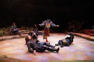 A performer with blue face paint and a costume adorned with colorful feather details stands center stage with arms outstretched over several other performers who appear to be falling asleep.