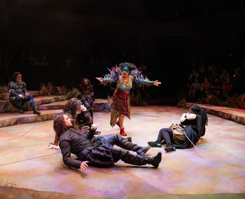 A performer with blue face paint and a costume adorned with colorful feather details stands center stage with arms outstretched over several other performers who appear to be falling asleep.