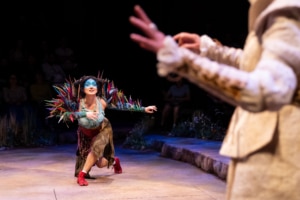 A performer with blue face paint and a costume adorned with colorful feather details kneels onstage and reaches toward another performer partially visible in the foreground.
